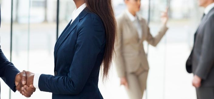 Handshake between a woman and a man, both in business attire, with two other people in suits discussing in the background